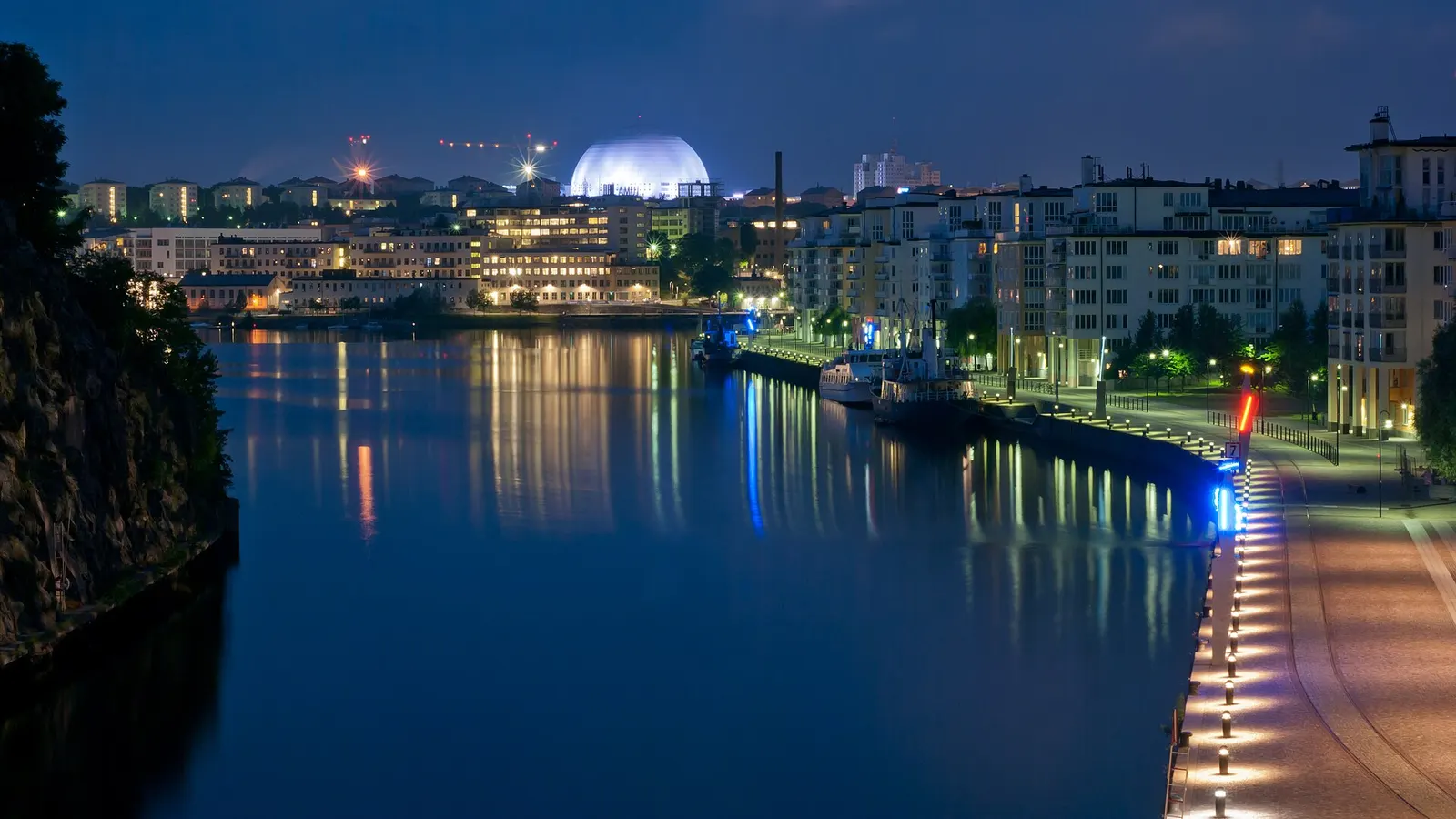 Flerbostadshus i Hammarby sjöstad i Stockholm en kväll, med ljusen speglade i kanalen och Globen i bakgrunden
