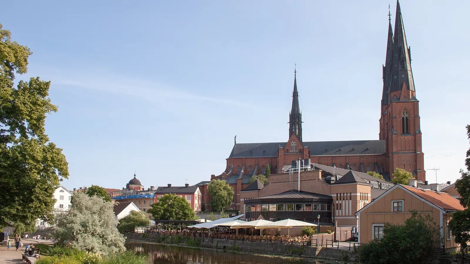 Uppsala domkyrka sedd från Fyrisån en sommardag, med äldre hus och uteserveringar längs ån